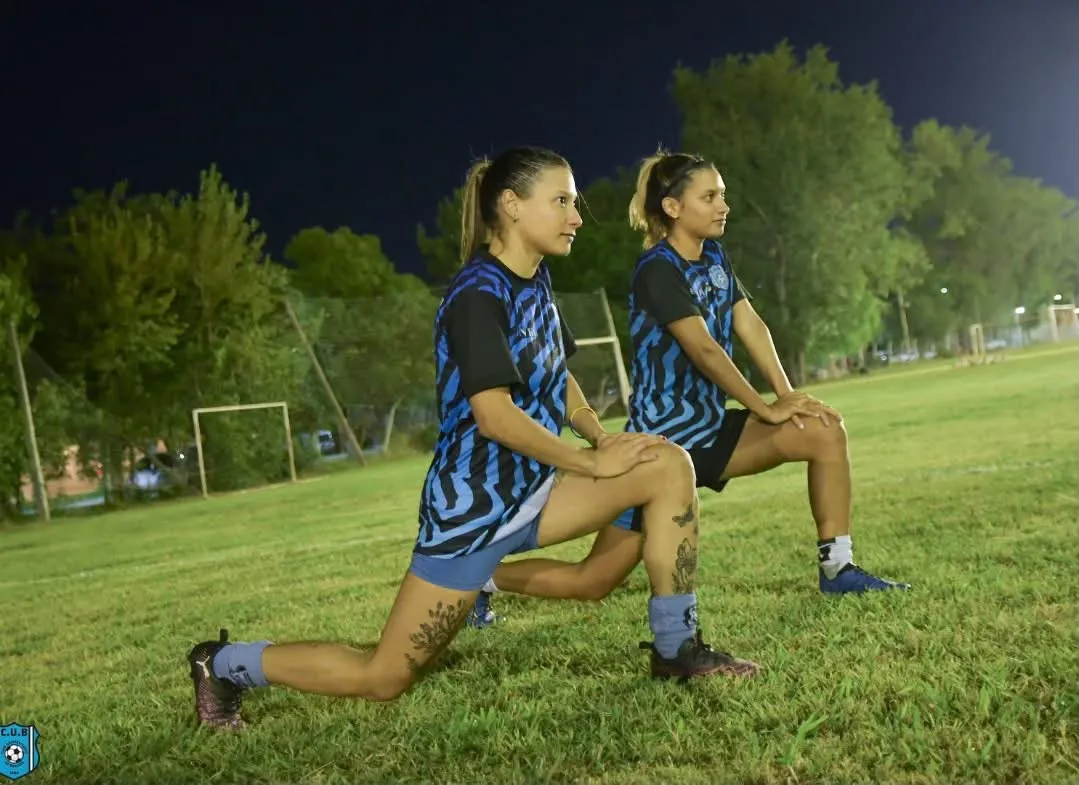 Dos jugadoras del plantel femenino elongando en la cancha durante un entrenamiento nocturno