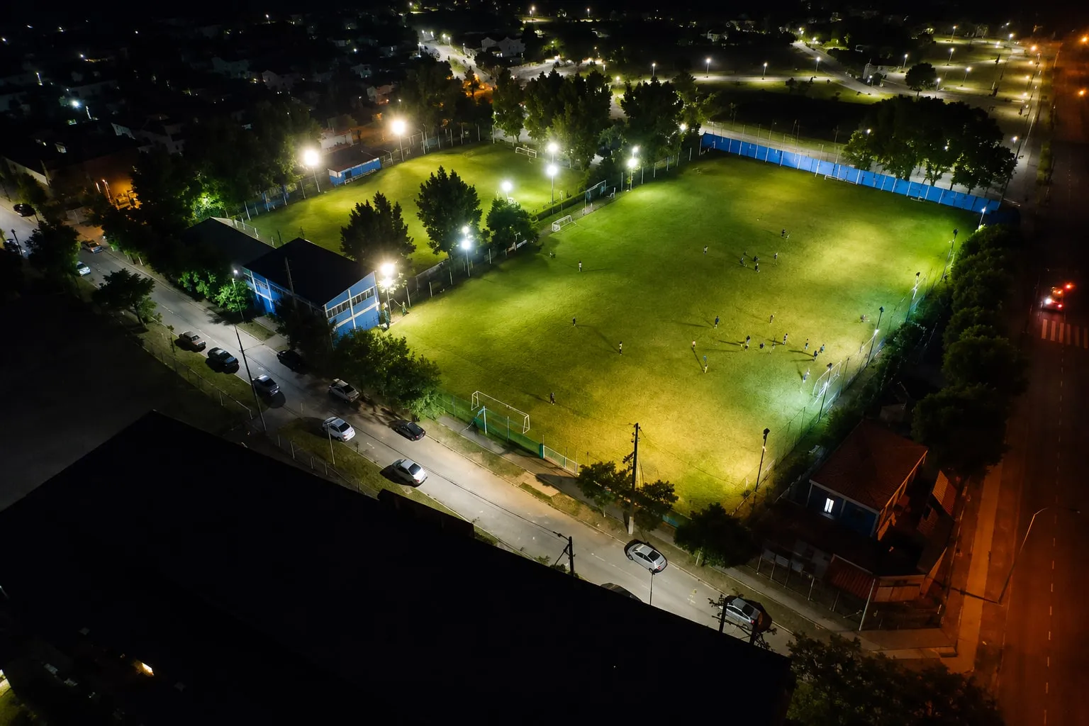 Vista aérea nocturna de la cancha del Club Universitario de Berisso con los reflectores encendidos, con un partido en juego, rodeada por las casas y las calles del barrio