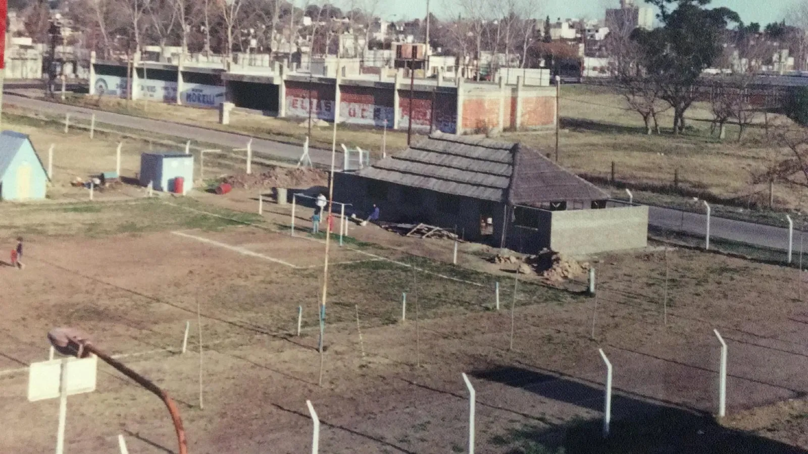 La cancha del club vista desde adentro, con el paredón perimetral y las casas del barrio detrás